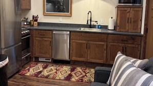 A compact kitchen with wooden cabinets, a stainless steel refrigerator, a dishwasher, and a microwave. There's a large mirror above the sink, a patterned rug on the floor, and a gray sofa with striped pillows. - Charming and Cozy Retreat in Newaygo, MI- The Rustic Barn dominium (Newaygo)