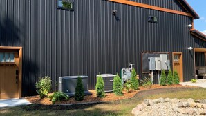 A large industrial building with a dark corrugated metal exterior and orange trim. There are two small windows and a door on the side. In front, there are outdoor air conditioning units, a small garden with shrubs, and a gravel area. - Charming and Cozy Retreat in Newaygo, MI- The Rustic Barn dominium (Newaygo)