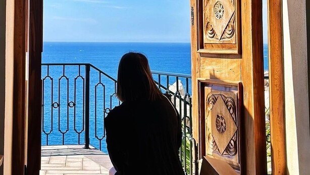 A view through an ornate wooden door with intricate carvings, leading to a balcony overlooking a clear blue sea.