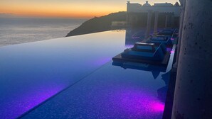An infinity pool with a view of the sunset over the ocean, a row of sun loungers, and a building with classical architecture in the background.