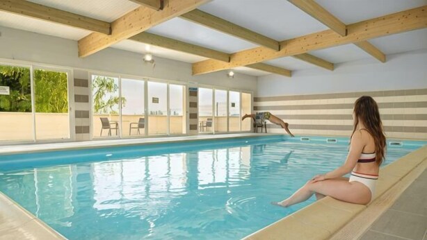 An indoor swimming pool with wooden beams, a tiled floor, and a person sitting by the pool. - Kyriad Sens (Sens)