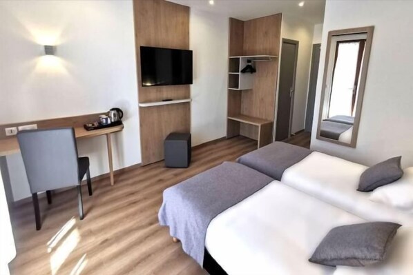 A modern hotel room with a wooden floor, a desk with a chair, a flat-screen TV, a wardrobe, and two beds. - Kyriad Sens (Sens)
