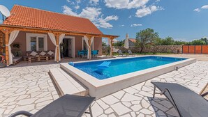 Un espace piscine doté d’une terrasse carrelée, de chaises longues et d’un coin salon couvert avec des coussins.