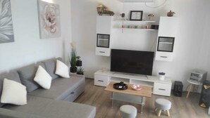 Un salon moderne avec un canapé gris, une table basse et deux poufs. Un téléviseur à écran plat est fixé au mur, au-dessus d’un meuble blanc doté d’étagères.