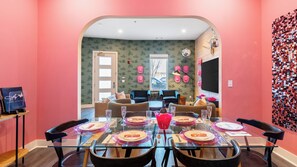 Zona pranzo con tavolo in vetro apparecchiato per sei persone, televisione e parete con decorazione a motivi.