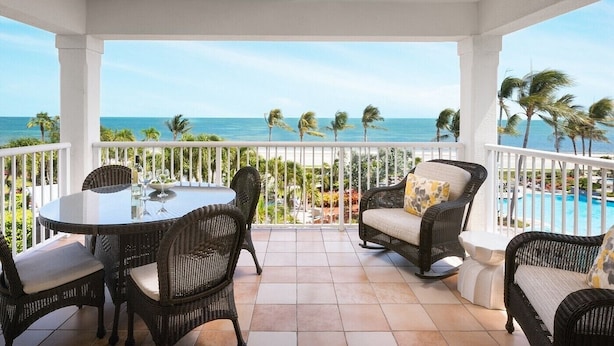 Een overdekt buitengebied met rieten meubels, een glazen tafel en uitzicht op de oceaan en palmbomen.