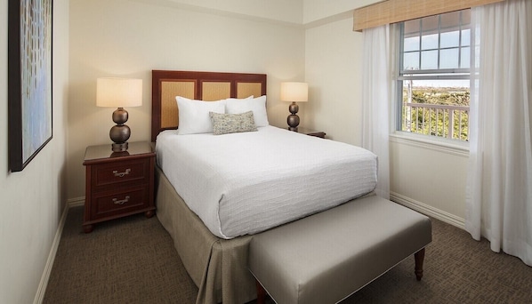A hotel room with a large bed, two bedside tables with lamps, a bench, a window with curtains, and a view of greenery outside.