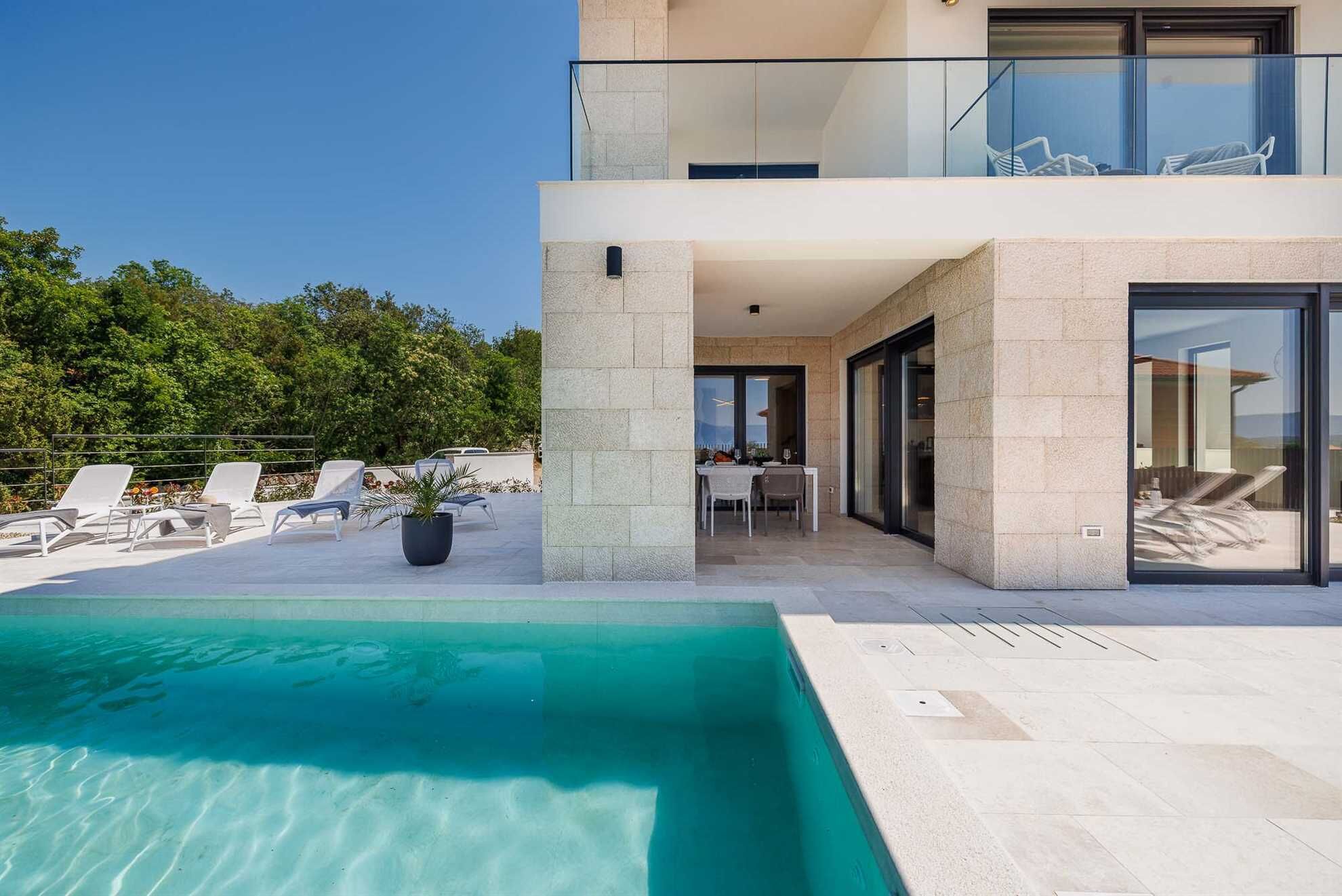 A modern two-story building with a swimming pool, outdoor seating, and a view of the surrounding greenery.