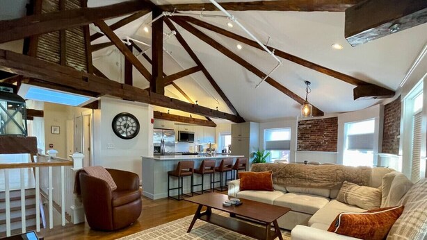 A spacious living room with a high ceiling, exposed wooden beams, a large sofa, a coffee table, and a kitchen area in the background. - 4 King Suites, Downtown, Outlets, Beach, Historic (Portsmouth)