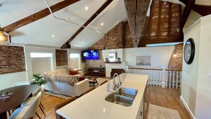 A modern kitchen with a large island, a dining area with a table and chairs, a living room with a sofa and TV, and a staircase leading to an upper level. - 4 King Suites, Downtown, Outlets, Beach, Historic (Portsmouth)