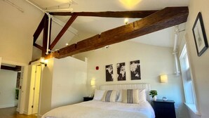 A bedroom with a large bed, wooden beams, a white headboard, and a small bedside table with a lamp. - 4 King Suites, Downtown, Outlets, Beach, Historic (Portsmouth)