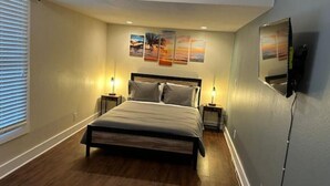 A bedroom with a bed, two bedside tables with lamps, a TV mounted on the wall, and artwork above the bed. - Sunny condo across the Bay in Pacific Beach! (San Diego)