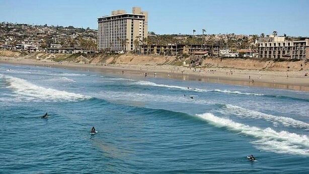 A coastal view with surfers in the water, a beach, and a hotel in the background. - Sunny condo across the Bay in Pacific Beach! (San Diego)