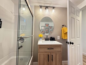 A modern bathroom with a glass shower enclosure, a wooden vanity with a sink, and a mirror with a decorative frame. - Modern 2Br + Sofa Bed + 2Bth in Miracle Mile (Los Angeles)