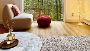 A modern living room with a large window, a comfortable sofa, a red pouf, and a decorative tree light.