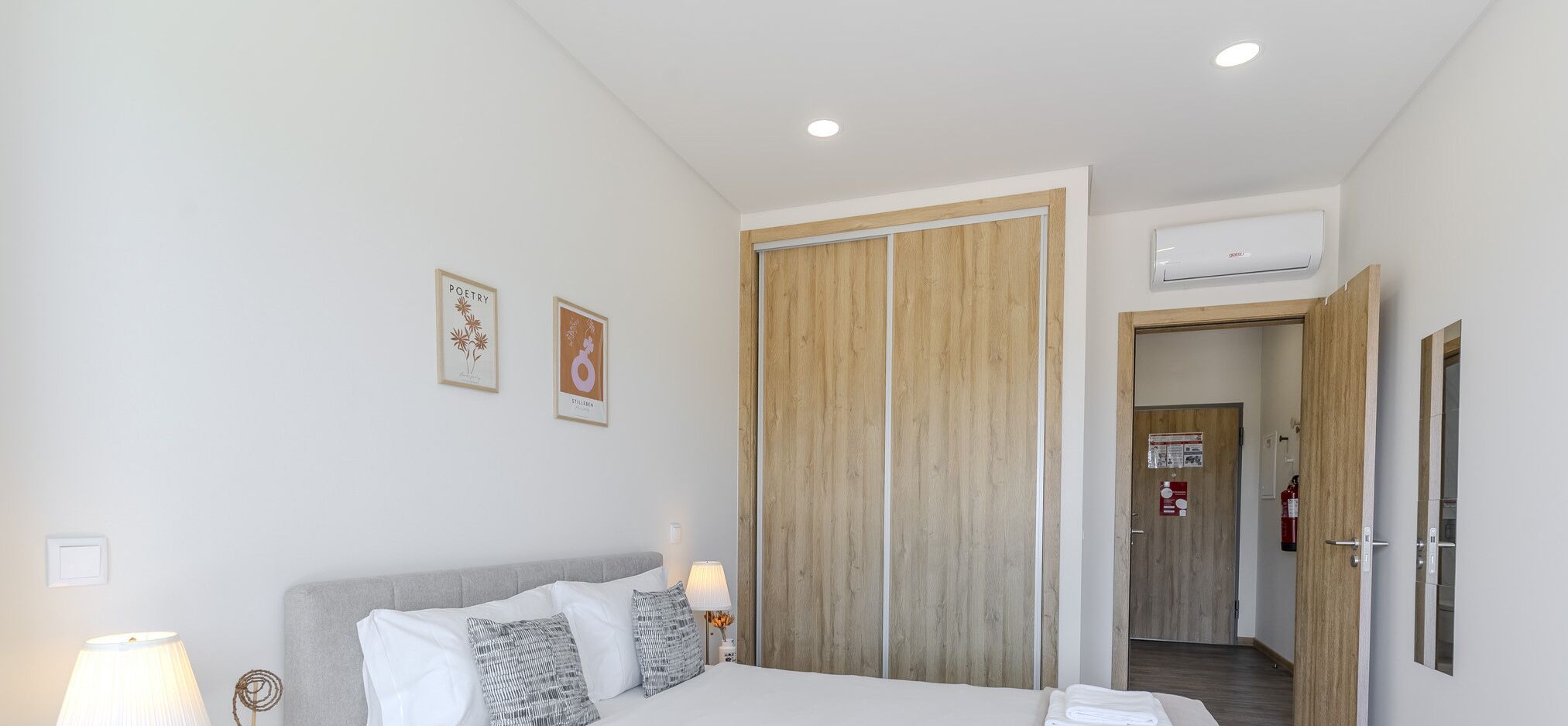 A hotel room with a bed, bedside lamps, a wooden wardrobe, and an air conditioning unit.