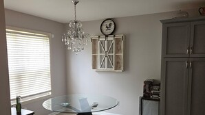 A dining area with a glass table, a chandelier, a rustic wall clock, and a wooden cabinet.