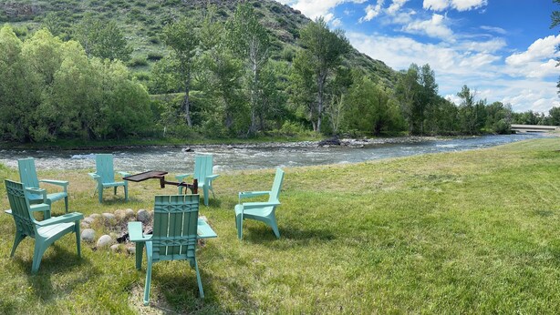 Entorno junto al río con mesas al aire libre y vistas a un río y colinas boscosas.