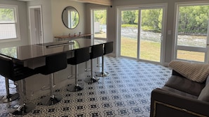Cocina moderna con una isla central, taburetes negros y amplias ventanas que ofrecen vistas a un río y follaje.