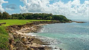 A coastal landscape with a grassy area, rocky shoreline, and a clear view of the sea.