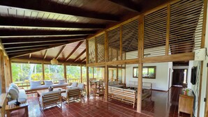 A spacious room with a tiled floor, wooden furniture, and large windows offering a view of greenery outside.