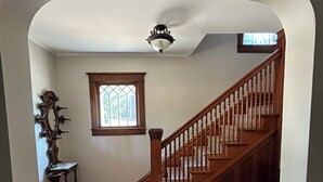 A wooden staircase with a handrail, a small table with a vase, and a window with a view of greenery. - The Bly House (Fulton)