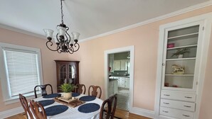 A dining room with a round table, chairs, a chandelier, a cabinet, and a kitchen in the background. - The Bly House (Fulton)