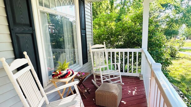 A covered porch with white rocking chairs and a wicker ottoman. - The Bly House (Fulton)