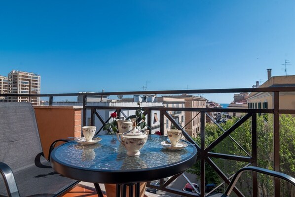 Una mesa redonda con superficie de vidrio y una silla, ubicada en un balcón con vistas a edificios y árboles.