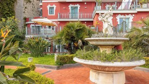 A courtyard with a fountain, lush greenery, and a red building with balconies. - Terrazza Aphrodite - Two Bedroom Apartment, Sleeps 5 (Salerno)