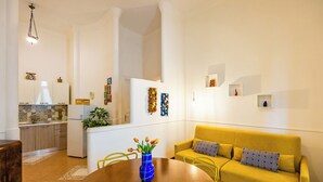 A modern living room with a yellow sofa, a wooden dining table with a vase of flowers, and a kitchen in the background.
