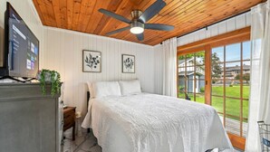 Un dormitorio con techo de madera, una cama con colcha blanca, ventilador de techo, televisor sobre un aparador y ventana con vista a una casa.