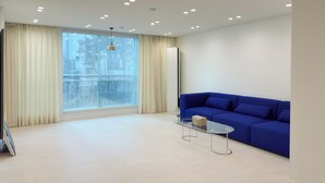 A modern living room with a blue sofa, a glass coffee table, and a large window with curtains.