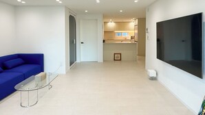 A modern living room with a blue sofa, a glass coffee table, and a kitchen area with a large TV.