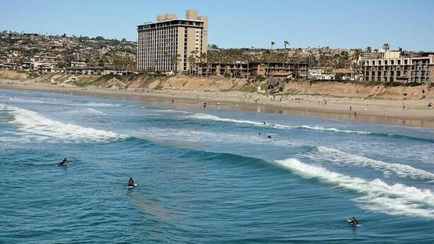A coastal view with surfers in the water, a beach, and a large hotel building in the background.