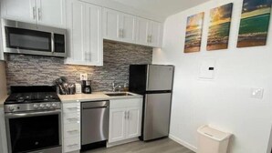 A modern kitchen with white cabinets, a stainless steel refrigerator, a built-in oven, and a microwave. There are three paintings on the wall and a trash bin.