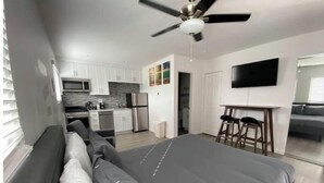 A modern bedroom with a large bed, a ceiling fan, a kitchen area with white cabinets, and a flat-screen TV mounted on the wall.