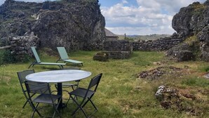 A round table with four chairs in a grassy outdoor area, surrounded by stone walls and rock formations under a partly cloudy sky. - Le P'tit Pelet, a house in the heart of the rocks (Fraissinet-de-Fourques)