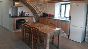A rustic kitchen with a stone wall, wooden ceiling beams, a dining table with chairs, a stove, a microwave, and a refrigerator. - Le P'tit Pelet, a house in the heart of the rocks (Fraissinet-de-Fourques)