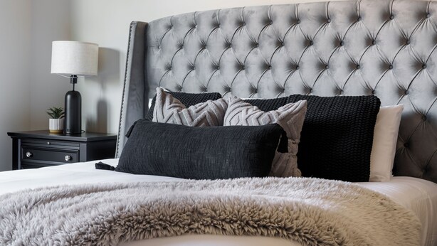 Camera da letto con testiera trapuntata, una soffice coperta beige e un comodino con lampada e una piccola pianta.