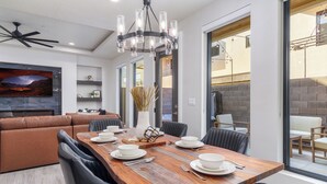 Een moderne eetruimte met een houten tafel voor zes personen, omringd door zwarte stoelen. Er is een plafondventilator, een kroonluchter en een grote televisie aan de muur bevestigd.