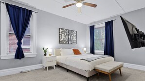 Un dormitorio con cama, mesitas de noche, ventilador de techo y ventana con cortinas.