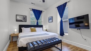 A bedroom with a bed, a bench, a TV, and two windows with curtains. - 5BR St. Louis Home | Backyard Fire Pit + Games (St. Louis)