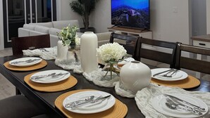Un comedor moderno con una mesa redonda dispuesta para seis personas, con platos blancos, cubiertos y jarrones decorativos. Un televisor grande está montado en la pared, y se ve una planta en maceta al fondo.