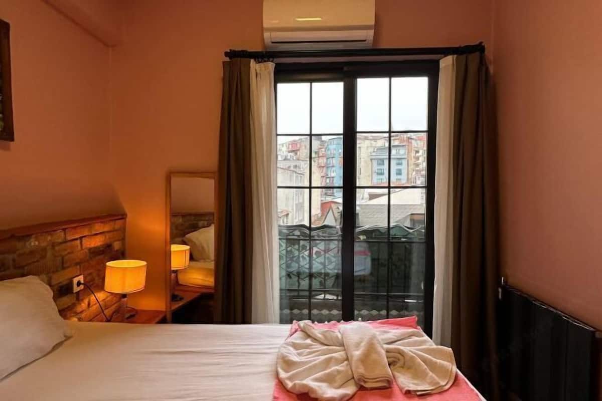 A hotel room with a bed, a nightstand with a lamp, a window with curtains, and a view of buildings outside.