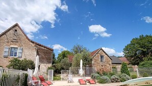 Un edificio de piedra con techo inclinado, un patio amurallado con sillones de descanso y una piscina.