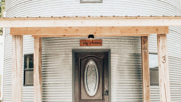 A corrugated metal building with a wooden porch, a door with an oval window, and a sign above the door. - Rustic Grain Bin Lodge with WiFi and AC in Loose Creek, MO (Loose Creek)
