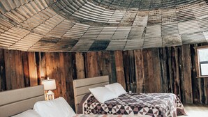 A rustic room with wooden walls and a corrugated metal ceiling. There are two beds with white and patterned bedding. - Rustic Grain Bin Lodge with WiFi and AC in Loose Creek, MO (Loose Creek)