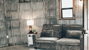 A room with a grey sofa, a small table with a lamp, and a window with a wooden frame. - Rustic Grain Bin Lodge with WiFi and AC in Loose Creek, MO (Loose Creek)