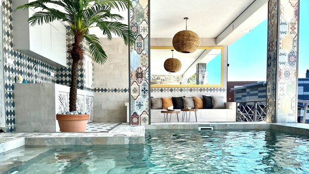 Ein Poolbereich mit gefliestem Boden, einer Palme und einer Sitzgelegenheit mit einer Couch und Tischen.